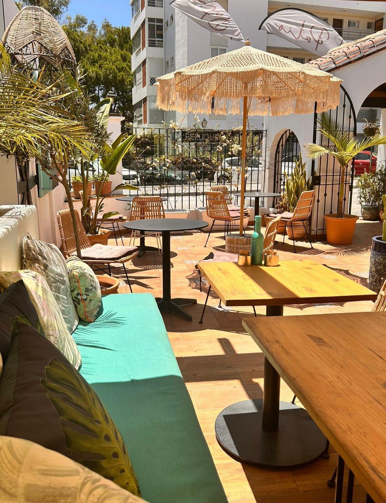 Outdoor café with wicker chairs, wooden tables, and a fringed umbrella under a sunny sky.